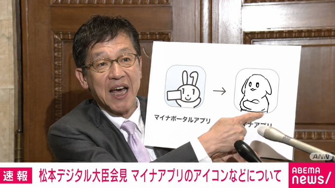 The image features a man wearing glasses and a suit holding up a sign with an illustration of a cartoon dog. The text on the sign reads "Change to the My Number Portal App Icon: Moving Away from Mina-chan news/livedoor.com/article/detail". This suggests that there is a change in the icon for the My Number portal app, and it has been decided to move away from the current design featuring a cartoon dog towards one resembling the My Number Card.