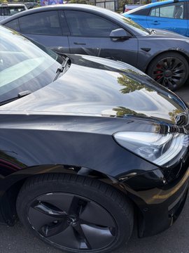 A close-up view of a black Tesla vehicle parked alongside a gray Tesla and a blue car, focusing on the front wheel and headlight.