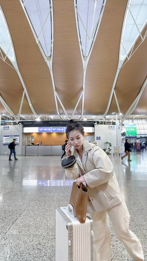 A young woman poses in an airport terminal with a silver suitcase and brown tote, her confident stance and stylish beige coat reflecting the polished but weary "dealing with weekend chaos" vibe of the post about navigating WEB3 liquidity issues.