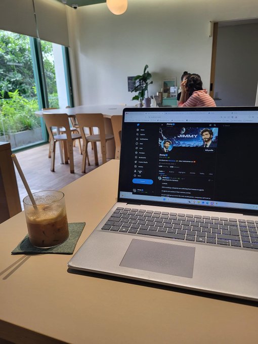 A laptop on a cafe table displays a Twitter (X) profile for "JIMMY" alongside an iced coffee, while a person sits at another table in the background — capturing a quiet moment of market reflection and narrative brainstorming that mirrors the post's "gm" tone of low trading volumes and thoughtful speculation about emerging crypto themes like robotics and AI agents.