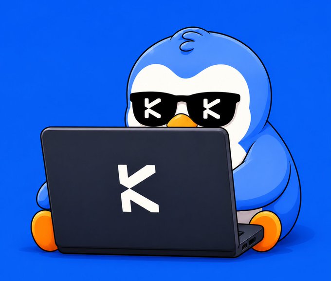A cartoon penguin with 'X' shaped sunglasses, a curly hair tuft, and orange feet, sitting behind an open laptop displaying a large 'K' logo on a solid blue background.
