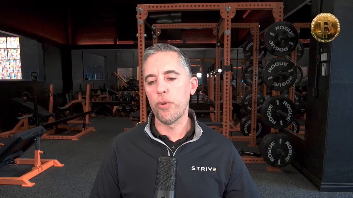 The image features a man wearing a black jacket, standing in front of a weight rack at a gym. He appears to be talking into a microphone or recording device while surrounded by various exercise equipment such as dumbbells and barbells. The background shows an indoor setting with a large window that allows natural light to enter the room.