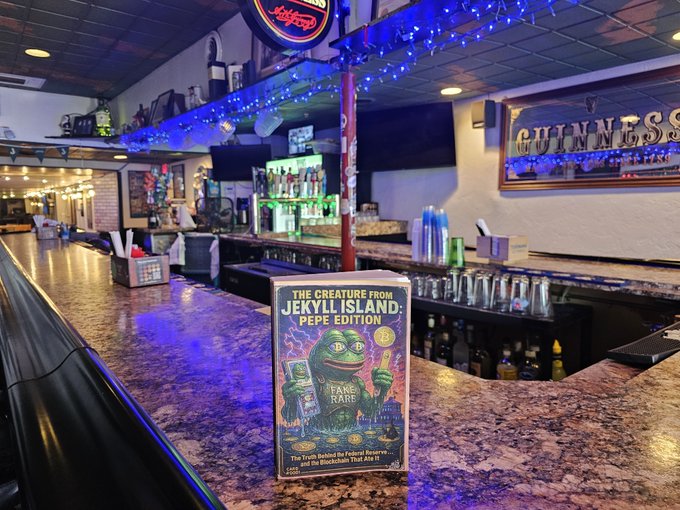 The same 'The Creature From Jekyll Island: Pepe Edition' parody trading card is displayed upright on a bar counter inside a pub with blue string lights and Guinness signage.