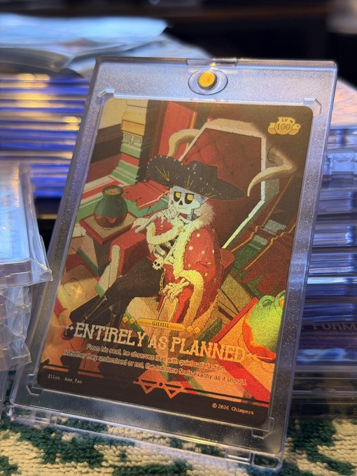 A holographic trading card titled 'ENTIRELY AS PLANNED' depicts the character Grillby from Undertale sitting in a chair, displayed in a protective plastic case.
