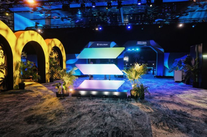 The image shows a large convention center with a stage featuring a colorful display of three-dimensional shapes, including a pyramid and a tree. The stage is surrounded by potted plants, creating an inviting atmosphere for attendees. In front of the stage, there are two potted plants placed on either side, adding to the overall aesthetic appeal of the event space.

The image also contains text that reads "The biggest reason @solana keeps attracting users isn't TPS It’s USER EXPERIENCE Crypto finally started feeling simple @phantom made wallets easy @Backpack introduced xNFTs @solflare simplified staking @Pumpfun made token launches instant Most chains focus on". This text suggests that the event is focused on promoting and discussing the new cryptocurrency, Solana. The presence of a stage with a colorful display indicates that it might be an exhibition or a launch event for the cryptocurrency.