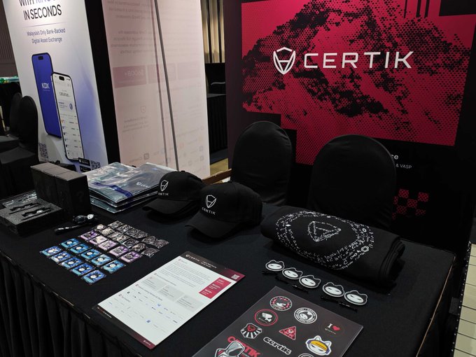 The CertiK team is set up at Booth D29 for the Malaysia Blockchain & Crypto Week event in Kuala Lumpur, with branded merchandise including black caps, a t-shirt, stickers, and informational materials displayed on their booth table. The backdrop prominently features the CertiK logo and name, reflecting the company's presence at this Web3 security and blockchain innovation conference.