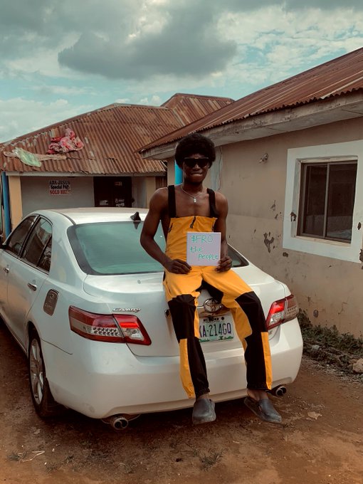 A man wearing a black tank top and yellow overalls sits on the trunk of a white Toyota Camry, holding a handwritten sign that reads "FRO the People." The license plate "AA-214GQ" and the rust-colored roof of a building in the background frame this grassroots promotion of the $FRO community token.