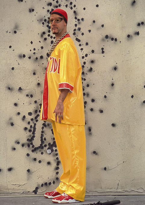 A man with glasses, a red bandana, and gold chains stands in profile wearing a matching yellow satin jersey and pants outfit with red accents, paired with red sneakers, against a weathered beige wall covered in dark spots.