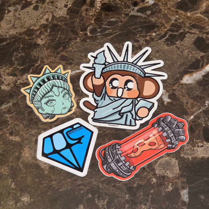 Four stickers are scattered on a brown marble surface, including a cartoon Statue of Liberty monkey, a classical bust of Liberty, a blue diamond shape, and a red pill-shaped sticker showing cartoon poop.
