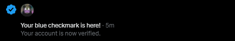 A Twitter notification reading 'Your blue checkmark is here! · 5m' and 'Your account is now verified.' with a verified badge and a profile picture of a person in a purple-lit room.