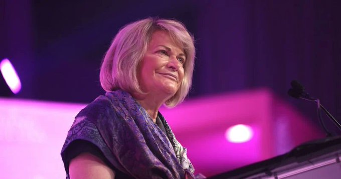 An older woman with shoulder-length blonde hair smiling warmly while standing behind a podium with a microphone, against a purple-lit background.