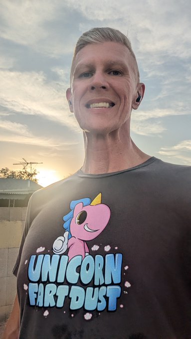 Selfie of a smiling man wearing a black t-shirt with a cartoon unicorn and the text "UNICORN FART DUST" against a sunset sky.