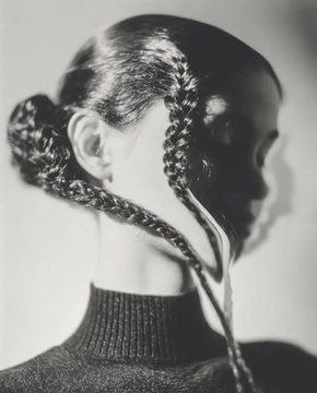 A black and white photograph shows a person from behind with elaborately braided hair, featuring small braids hanging down by their neck while wearing a dark turtleneck sweater.