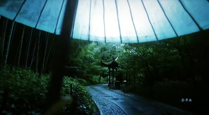 A view from beneath an umbrella looking down a wet stone path through lush greenery toward a pavilion in the distance, with Chinese characters '金泽雨' in the corner.