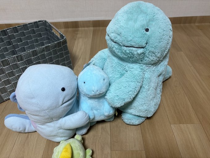 Three plush dolphin toys in varying shades of blue and teal sit on a wooden floor next to a gray checkered storage basket.