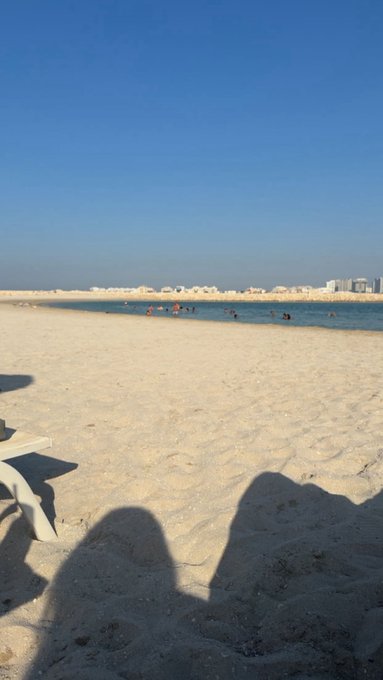 A beach scene with light tan sand, calm turquoise water, and distant buildings under a clear blue sky, with shadows visible in the foreground.