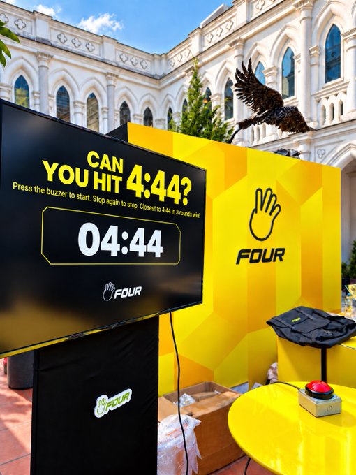A bright yellow outdoor booth invites passersby to test their timing skills with a giant red buzzer and digital display reading "CAN YOU HIT 4:44?" alongside the "FOUR" meme coin logo, set against an ornate white Gothic-style building. The setup matches the challenge described, offering three attempts to stop a timer as close to 4.44 seconds as possible to win an iPhone 18 Pro.