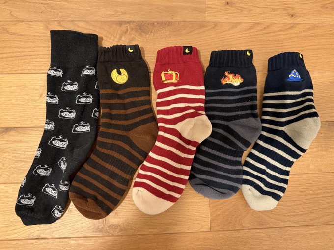 Five pairs of patterned crew socks are laid out in a row on a wooden floor, each featuring different designs including small cars, stripes, a TV, a flame, and a UFO.