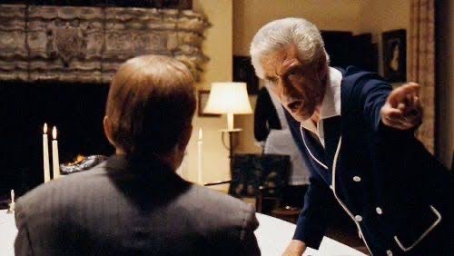 A film still shows an older white-haired man in a dark jacket angrily pointing at a seated person across a table in a warmly lit room.