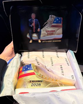 The image shows a person holding an open box of limited edition "$TRUMP 2028" sneakers, featuring an American flag design and the slogan "LET'S KEEP WINNING!" on the inside lid, alongside a figurine of Donald Trump. This aligns with the post's discussion of $TRUMP co-founder Bill Zanker teasing expansions of the Trump brand into entertainment ventures like a mobile game, TV show, and amusement park.