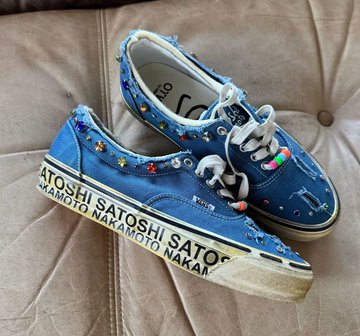 A pair of blue OTW by Vans × Satoshi Nakamoto "Era 95" sneakers featuring colorful lucky charm embellishments and "SATOSHI SATO" text along the sole stripe, photographed on a tufted cushion.