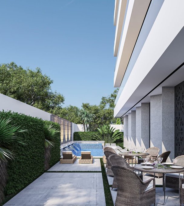 A modern luxury outdoor patio features wicker dining furniture, a small swimming pool, manicured hedges and palm trees, with sleek white architectural overhangs against a clear blue sky.