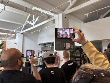 The image shows a group of people gathered around, with one person holding up their phone displaying a video or photo. There are several other individuals present as well, some standing closer together while others are more spread out across the scene. The focus on what is in the image and how it relates to the post suggests that this gathering might be related to an event or occasion where people have come together to watch or share content on their phones.

The presence of multiple people with their phones indicates a social setting, possibly at a cafe or similar venue. The group's interest in capturing moments through photos and videos further supports the idea of a shared experience among friends or acquaintances.