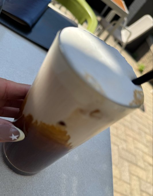 A hand holding a nearly empty caramel macchiato-style iced coffee in a clear plastic cup, showing some smudges inside the cup, on a balcony or patio setting.
