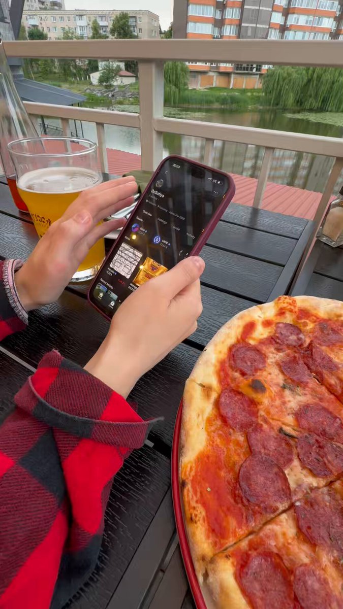 The image shows a person sitting at an outdoor table with a pizza in front of them. The person is holding a cell phone up to their face, possibly taking a selfie or looking at something on the screen. There are also two glasses on the table - one containing beer and another filled with water.