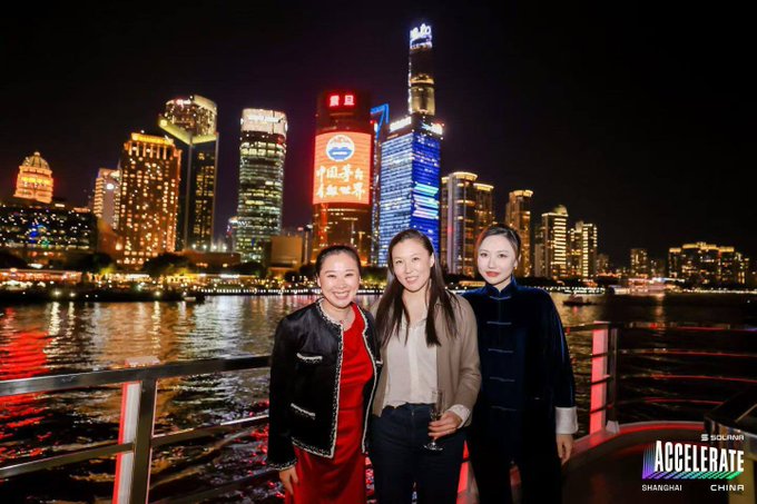 Three women pose for a photo at night in Shanghai, China, with the illuminated Pudong skyline and river in the background; a 'SOLUNA ACCELERATE SHANGHAI CHINA' logo appears in the corner.