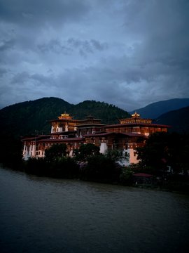 A majestic Bhutanese dzong (monastery-fortress) glows warmly in the twilight, perched along a riverbank with misty mountains rising behind it under a dramatic, overcast sky. The illuminated traditional architecture with its distinctive tiered roofs and white walls exemplifies the timeless cultural and spiritual character that defines Bhutan.