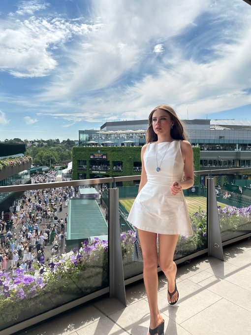 A woman in a white sleeveless dress and pendant necklace stands on a balcony overlooking a tennis venue, likely Wimbledon.