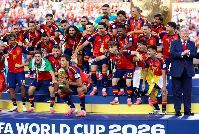 The Spain national football team celebrates with the World Cup trophy on a podium marked 'FIFA World Cup 2026', joined by a man in a suit.