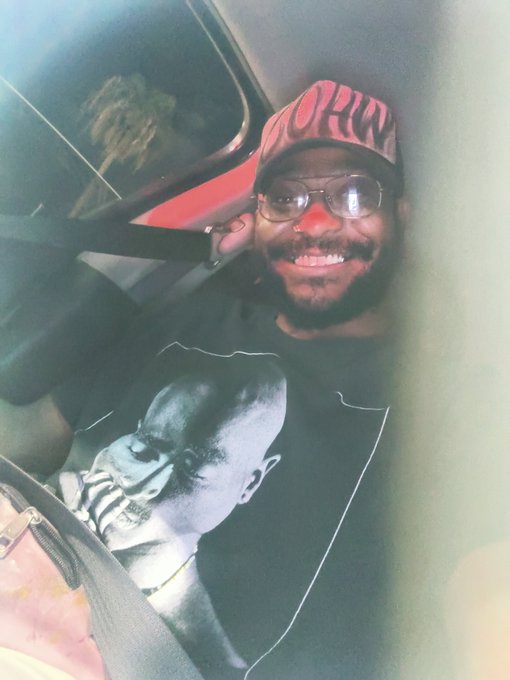 A man wearing glasses, a pink and black 'BORN' cap, and a black graphic t-shirt smiles while sitting in a car.
