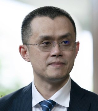 A man in a dark suit, white shirt, and blue striped tie wears wire-rimmed glasses in a close-up portrait, with a blurred background suggesting a formal setting. The image appears to accompany news about Binance CEO Changpeng Zhao (CZ) commenting on EU regulatory actions regarding a MiCA license.