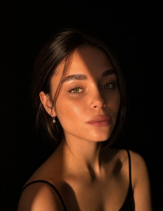 A portrait of a woman with dark hair and defined brows, illuminated by dramatic warm lighting against a black background, wearing a black spaghetti strap top.