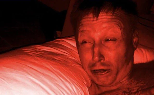 A close-up photograph of a man in a dimly lit setting with a reddish tint, looking at the camera with his mouth slightly open.