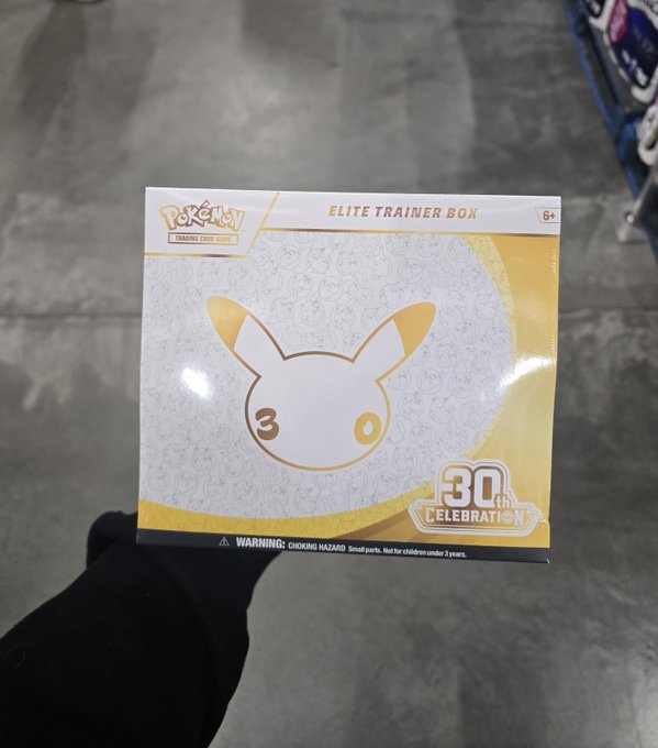 A hand holds up a sealed Pokémon Trading Card Game Elite Trainer Box featuring a gold and white design with a large Pokéball containing the number '30', commemorating the 30th Celebration, with a 'WARNING: CHOKING HAZARD' label visible at the bottom.