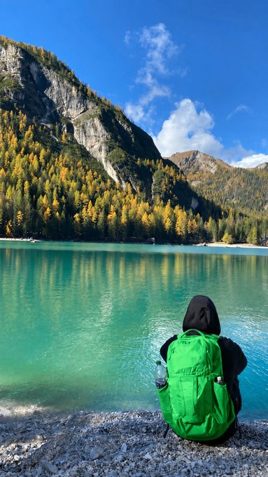 A person wearing a green backpack and black hoodie sits on a rocky shore, looking out at a vibrant turquoise lake surrounded by autumn-colored trees and rocky mountains.