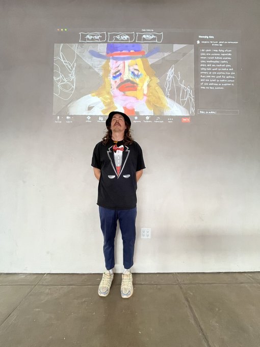 A man wearing a black t-shirt with a tuxedo graphic, blue jeans, and a bucket hat, standing in front of a wall projecting an AI-generated cartoon portrait of himself alongside a chat interface.