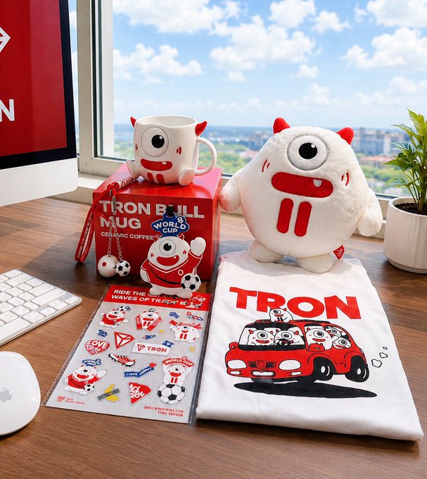A product display featuring TRON BULL merchandise including a ceramic coffee mug, plush toy, sticker sheet, and a t-shirt graphic showing characters in a red car, set on a wooden desk by a window.