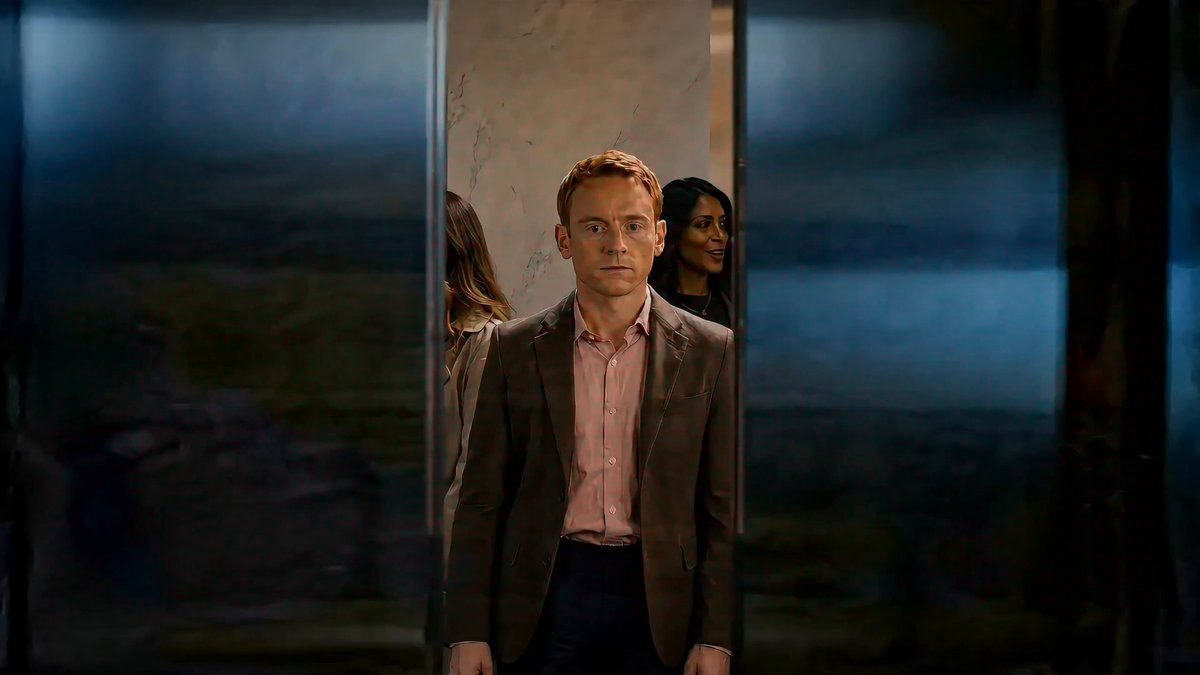 The image features a man wearing a suit standing in front of a glass door with other people visible through the glass as well. The scene appears to be set in an elevator, possibly at a movie theater or a similar venue where people gather for events. The main focus is on the man in the suit and his surroundings, creating a sense of anticipation and excitement related to the event taking place.