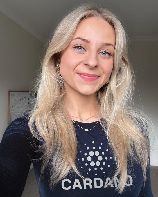 A selfie of a smiling blonde woman wearing a navy blue Cardano (CARDANO) shirt and gold jewelry.