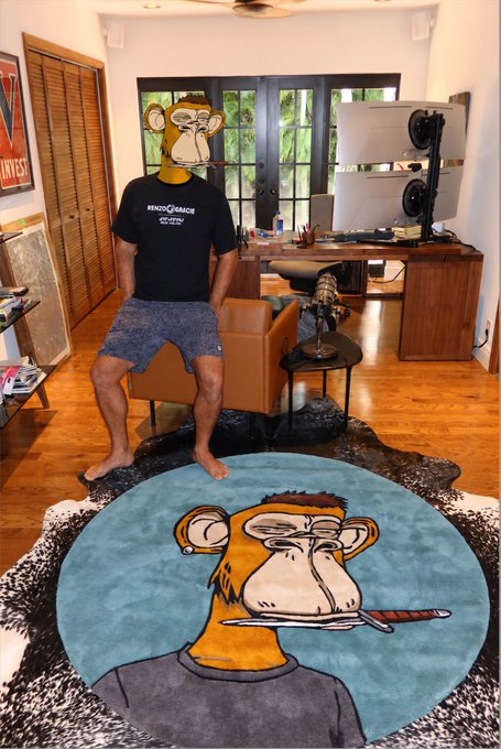 A photo of a person wearing a Bored Ape Yacht Club-style cartoon monkey mask standing in a living room, with a matching ape-themed rug featuring the same character smoking a cigar on the floor.