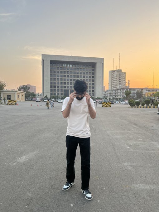 A young man in a white t-shirt and dark pants stands with his hands covering his face in a large paved lot with buildings in the background at sunset.