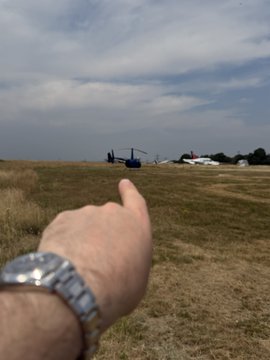 A first-person view of a hand pointing toward two helicopters on a grassy airfield under a partly cloudy sky, with a silver wristwatch visible on the wrist.