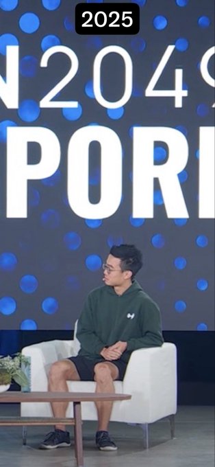 The image shows a man wearing a green hoodie and shorts sitting on a white couch on stage at an event with "2025" and "SPORTS" partially visible on a blue dotted backdrop. The "lock in" caption humorously reflects how he's giving a professional presentation while dressed casually, prioritizing the content over his appearance.