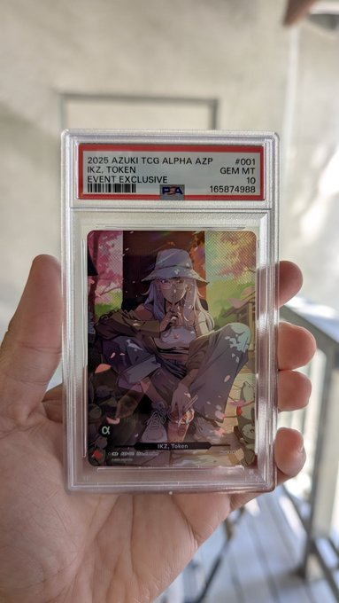 A hand holds a PSA-graded trading card in a protective case, labeled '2025 AZUKI TCG ALPHA AZP #001 IK.Z. TOKEN EVENT EXCLUSIVE GEM MT 10' with serial number 165874988B, depicting an anime character with lavender hair.