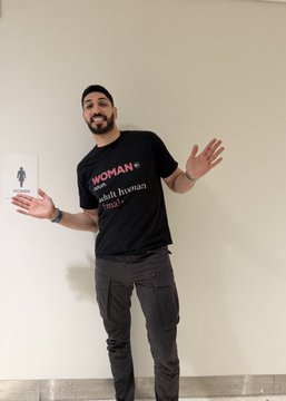 Photo of a smiling man wearing a black 'WOMAN = adult human ♀' t-shirt, gesturing with his hands while standing against a white wall.