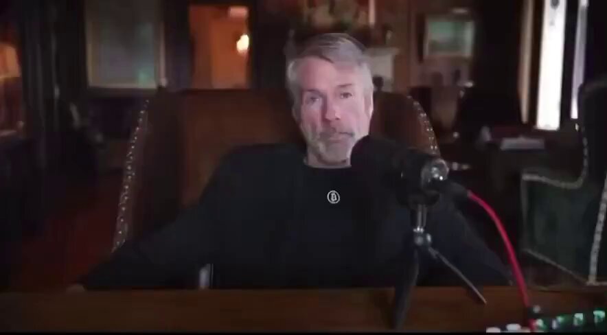 The image features an interview with Michael Saylor who is seated at a wooden desk surrounded by books and various objects. He appears to be looking directly into the camera, engaging with the viewer. The backdrop suggests a cozy room setting with a chair and couch nearby. In the foreground of the photo, there's a microphone close to him, indicating that this might be an interview or discussion on topics related to Bitcoin or digital currency.